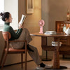 A woman sits reading a book in a modern interior while a pink portable rechargeable handheld fan rests on the table.
