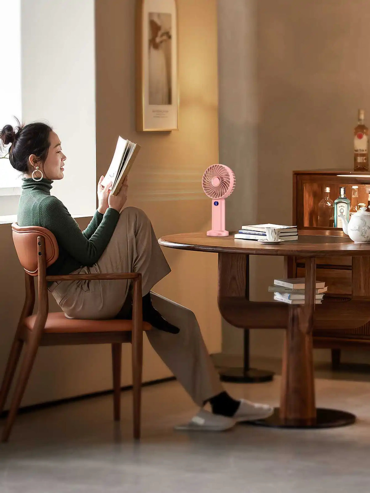 A woman sits reading a book in a modern interior while a pink portable rechargeable handheld fan rests on the table.