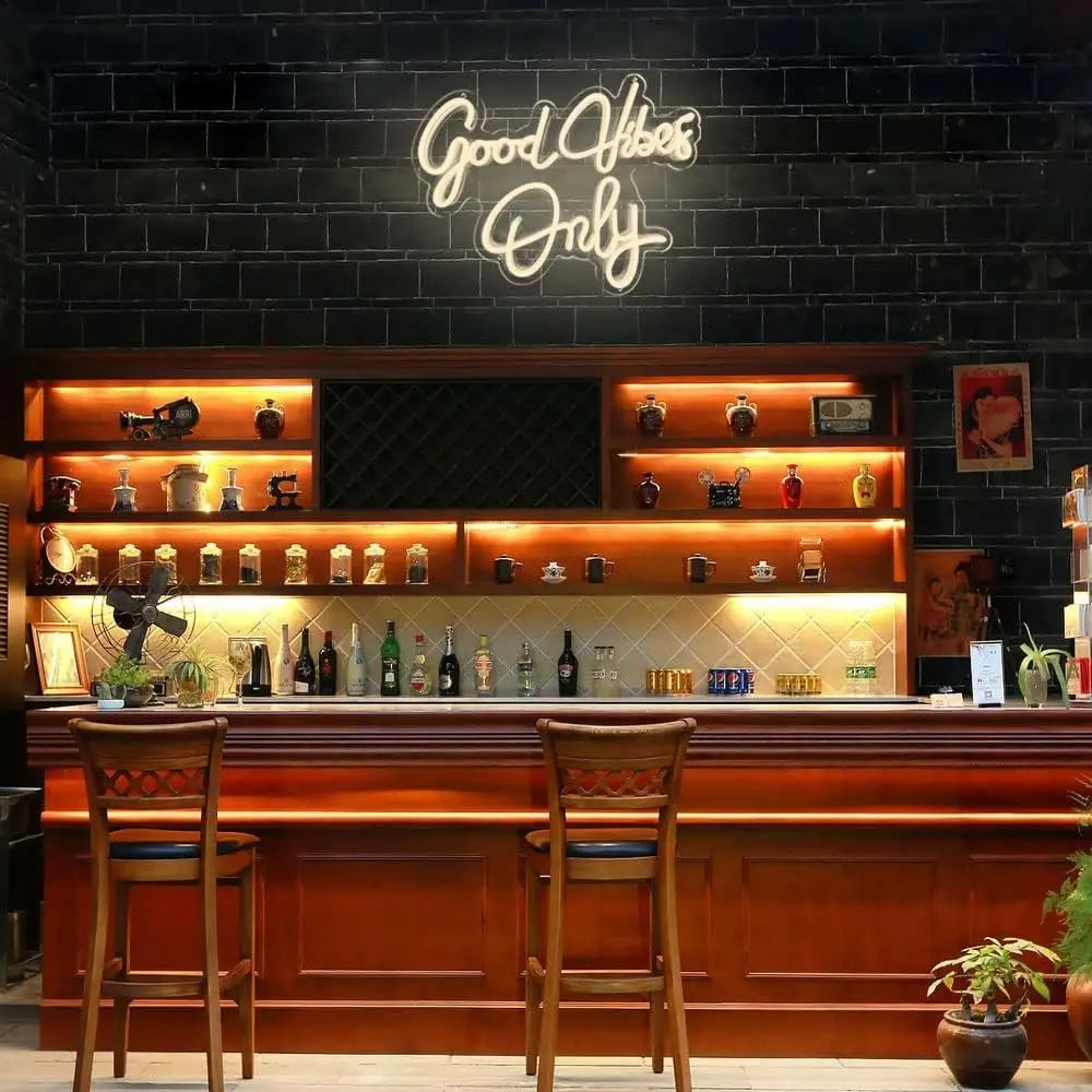 A bar counter with a wooden top and bar stools in a dimly lit interior featuring a neon sign that reads Good Vibes Only