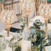 A decorative crystal candle holder with a golden finish is displayed among greenery and white flowers.