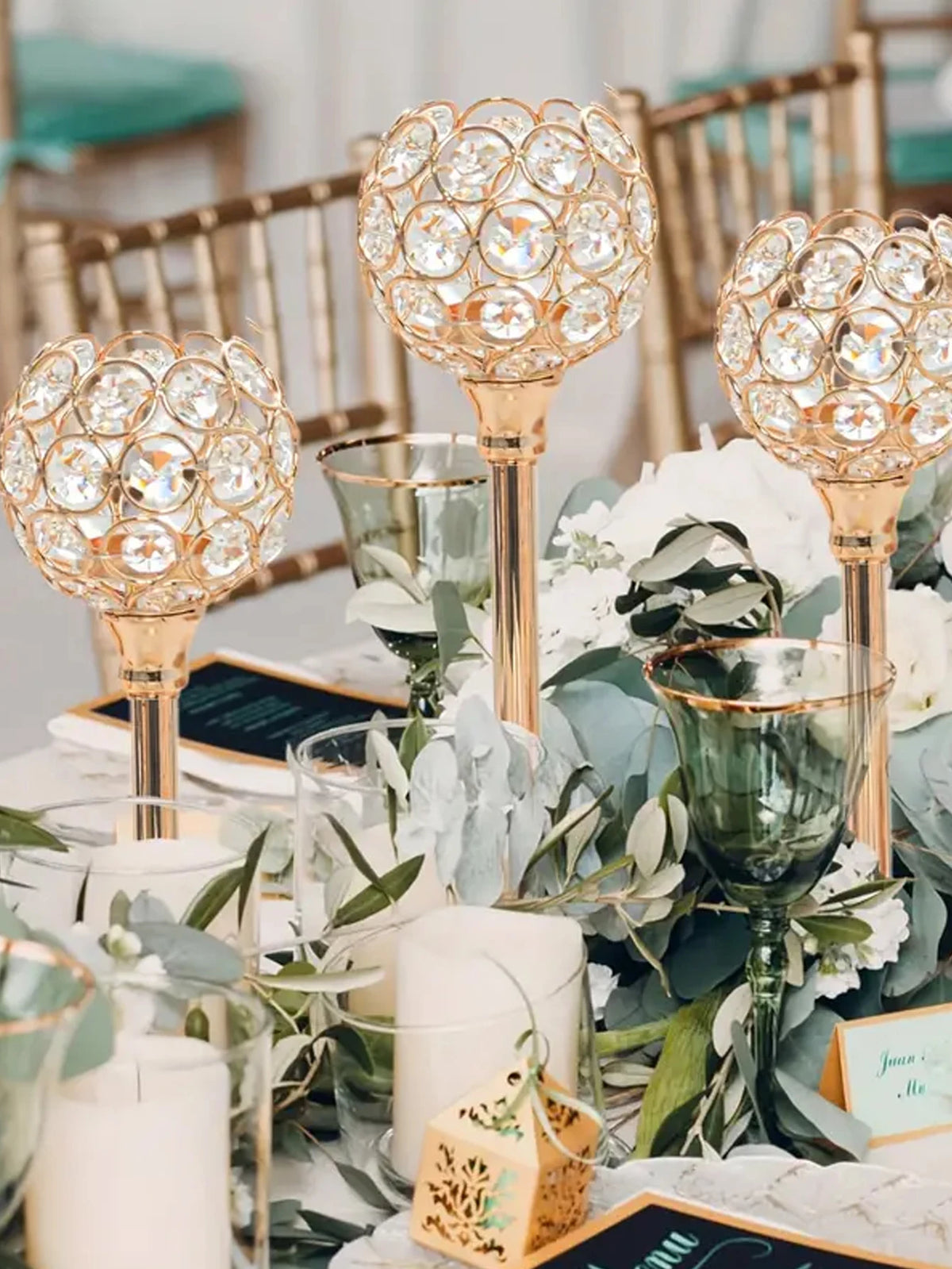 A decorative crystal candle holder with a golden finish is displayed among greenery and white flowers.