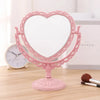 Espejo de maquillaje con forma de corazón rosa de doble cara y marco ornamentado sobre una superficie de madera.