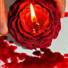 Eine Hand hält eine tiefrote Rosenkerze mit brennender Flamme, die Wachs auf eine weiße Oberfläche tropft.
