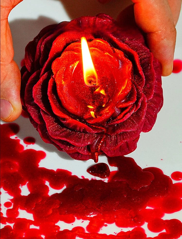 Eine Hand hält eine tiefrote Rosenkerze mit brennender Flamme, die Wachs auf eine weiße Oberfläche tropft.