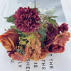A bouquet of artificial mixed flowers featuring deep red roses, orange daisies, and textured chrysanthemums.