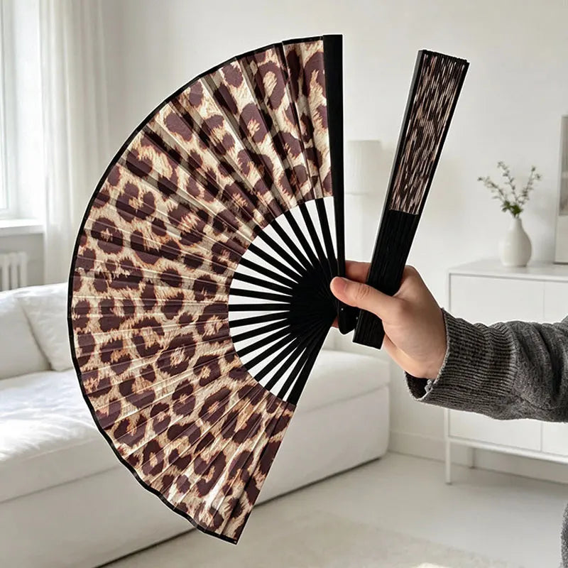 Eine Hand öffnet einen faltbaren, handlichen Ventilator mit Leopardenmuster in dunkelbraunen und schwarzen Tönen.