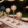 Three gold metal glass candle holders arranged on a wooden table with white lace tablecloth and silverware.