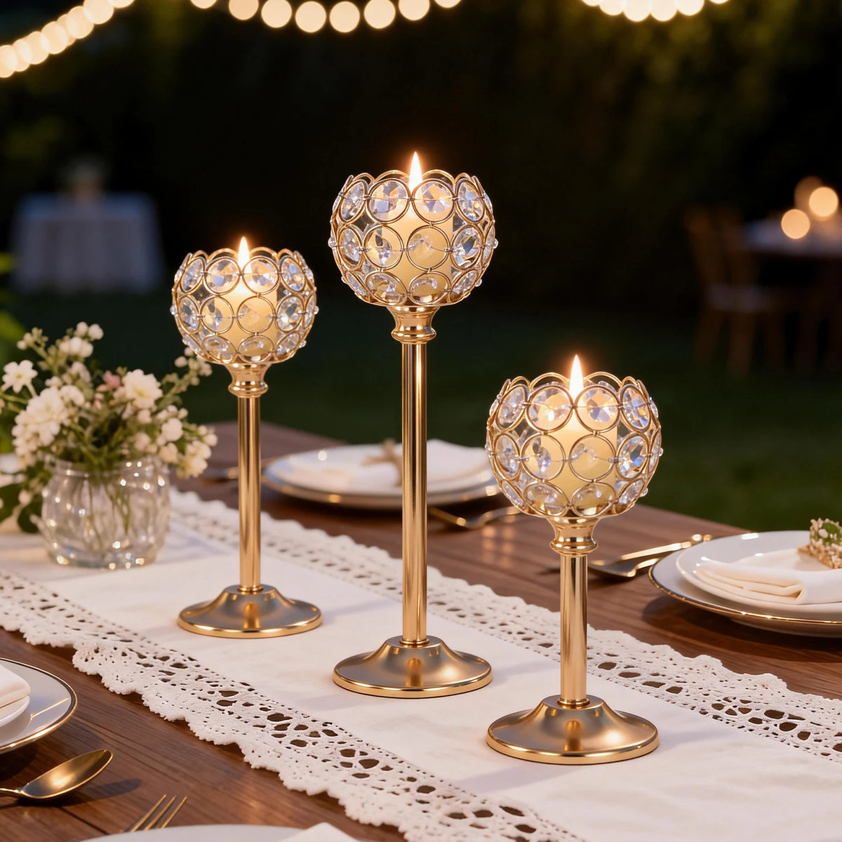 Three gold metal glass candle holders arranged on a wooden table with white lace tablecloth and silverware.