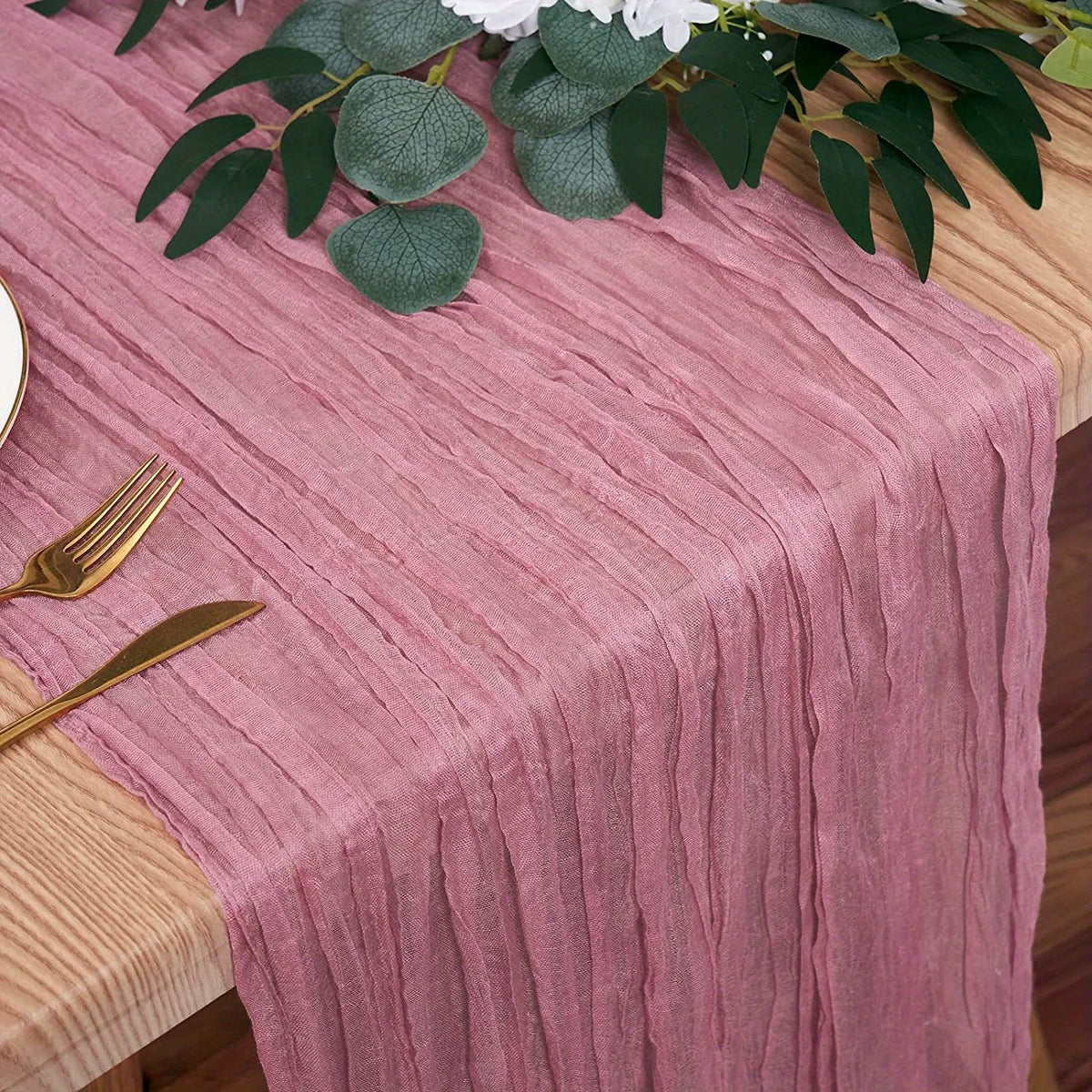 A pink linen table runner with vertical pleats is set on a wooden table, featuring gold cutlery and green foliage.