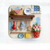 A white ceramic tray holds various kitchen items like wooden spoons, a woven basket, and a potted pink tulip plant.