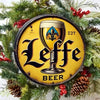 A vintage metal tin sign for Leffe Beer with a black silhouette of a beer glass on a yellow background, decorated with evergr