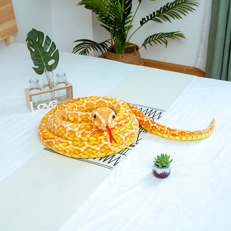 An orange and yellow giant realistic snake plush toy coiled on a white table with potted plants in the background.