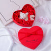 A red heart-shaped box filled with rose flowers and a white bear soap, resting on a white fabric.
