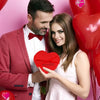 A man and a woman smiling while holding a red heart-shaped gift box with a plush bear inside.