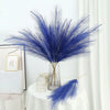 A tall glass vase holds a vibrant blue artificial pampas grass bouquet on a white draped table.