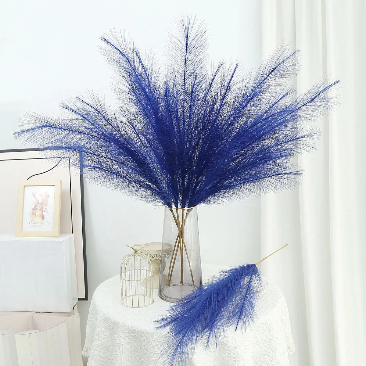 A tall glass vase holds a vibrant blue artificial pampas grass bouquet on a white draped table.