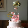 A pink birthday cake decorated with frosting swirls, topped by a gold glitter balloon and a pink flamingo candle shaped like 
