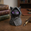 A dark gray gothic hooded cat statue with glowing purple eyes sits on a wooden surface next to an antique pen and stacked boo