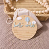 A wooden birth announcement sign with clouds and a hot air balloon design resting on a beige fabric.