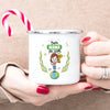 A hand holds a white enamel mug with a green design featuring a globe and laurel leaves, decorated with the text Best Mama de