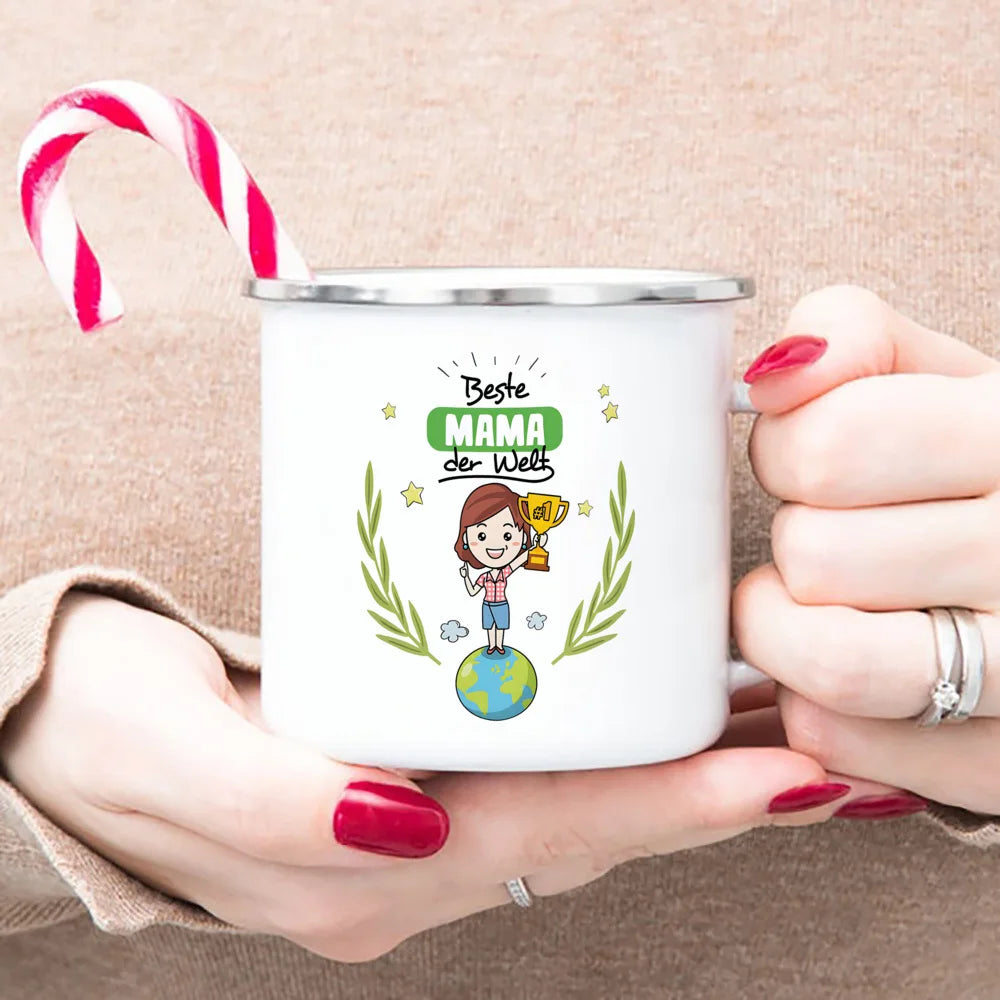 A hand holds a white enamel mug with a green design featuring a globe and laurel leaves, decorated with the text Best Mama de