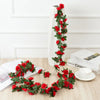 A long garland of red silk roses with green leaves displayed against a white wall and on a wooden surface.