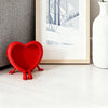 A bright red heart-shaped 3D photo frame ornament resting on a white wooden surface next to a framed picture.
