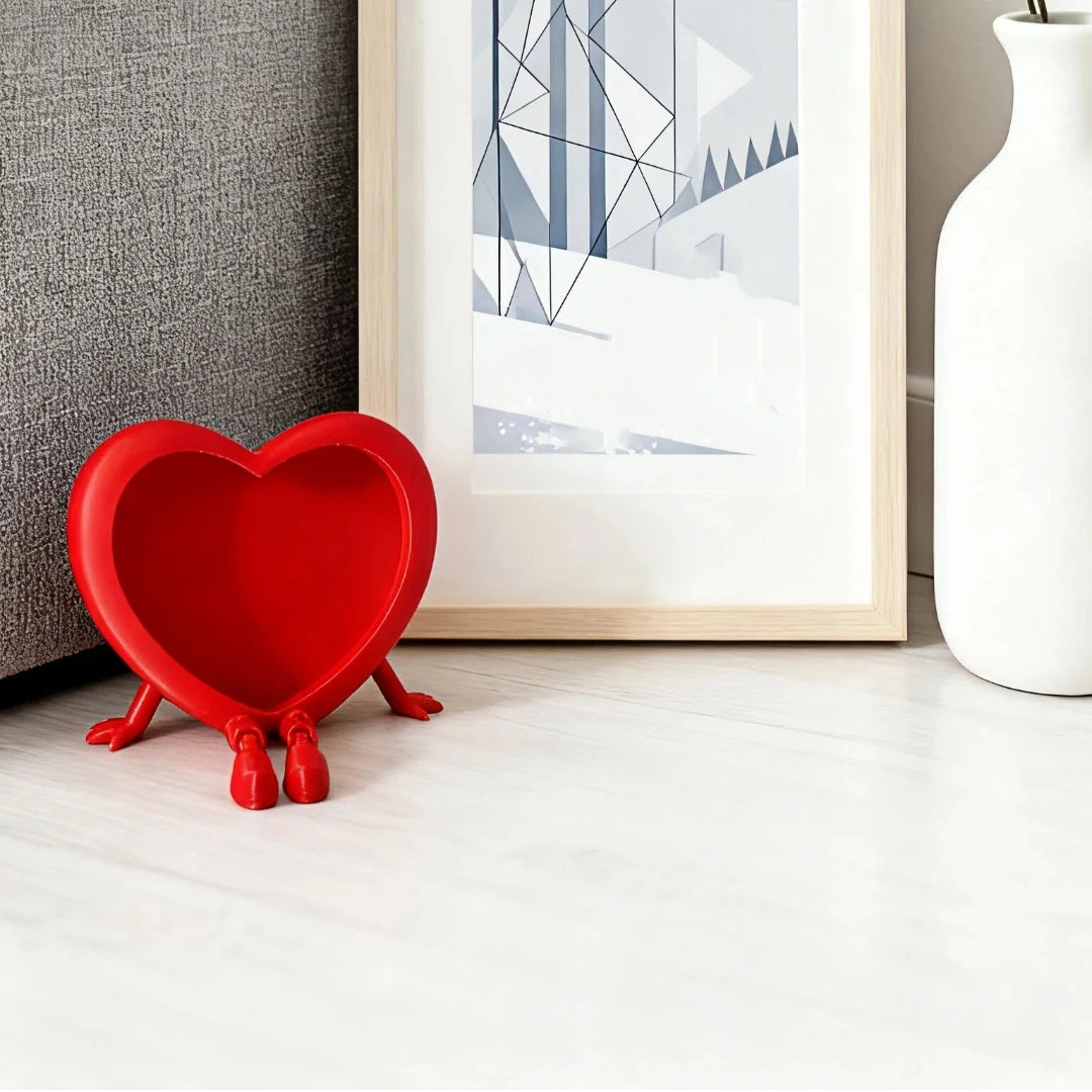 A bright red heart-shaped 3D photo frame ornament resting on a white wooden surface next to a framed picture.