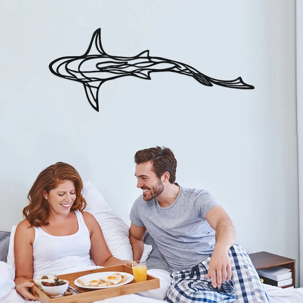 A couple is sitting on a bed in a bright room, looking at a black line sculpture of a shark on the wall.