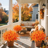 Two potted artificial fall foliage plants decorate a porch railing in front of a house with autumn trees.