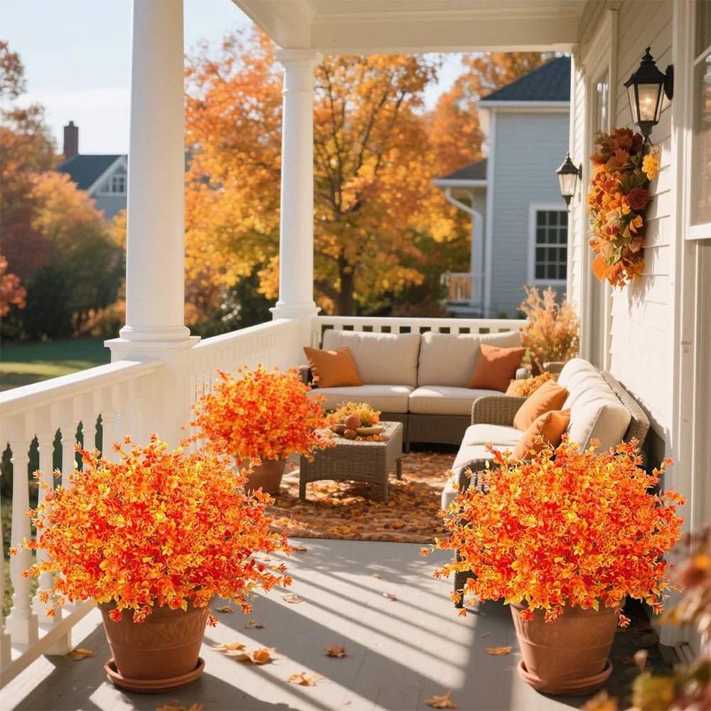 Two potted artificial fall foliage plants decorate a porch railing in front of a house with autumn trees.
