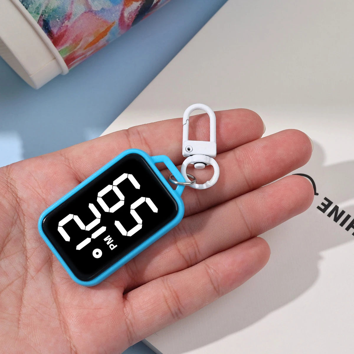A hand holds a blue LED keychain pocket watch with a digital display showing 2:00 PM.