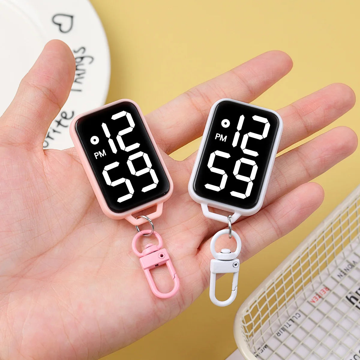 A hand holds a pink LED keychain pocket watch with a digital display showing 12:59 PM.