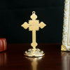 A golden metal Jesus cross ornament with a decorative base, displayed on a dark wooden surface.