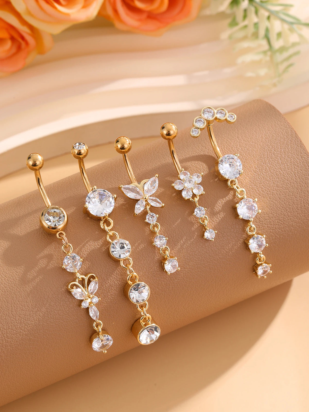 A set of five gold tone butterfly and flower belly rings displayed on a beige surface with soft floral accents.