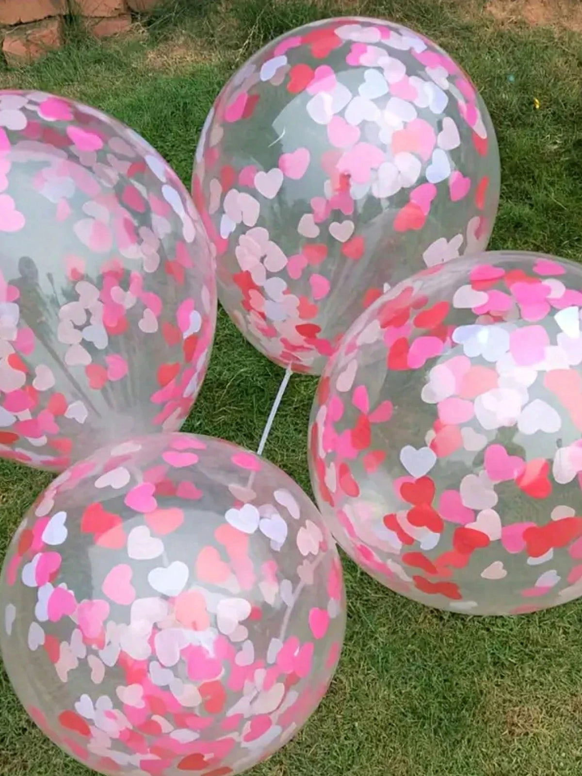 A collection of clear balloons filled with pink and red heart-shaped sequins confetti scattered on green grass.
