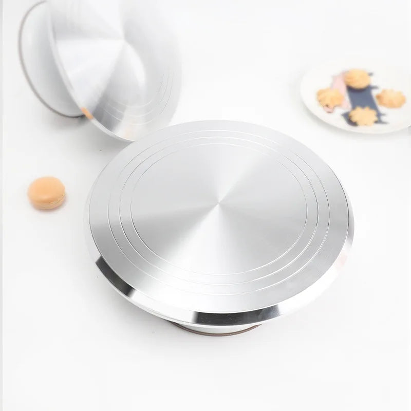 A silver aluminum cake turntable with a rotating platform and a small light-colored object nearby.