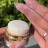 A hand holds a glass jar of Fairy Body Glitter Highlight Powder with a gold lid and white product, showing glitter on the fin