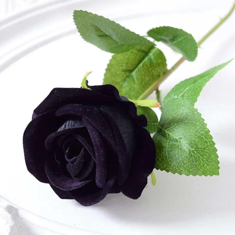 A deep black velvet artificial rose with a green leaf on a white background.