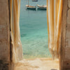A view through a window with yellow striped curtains showing a turquoise sea and boats.