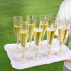 A set of clear plastic champagne flutes with glitter detailing displayed on a white platter outdoors.