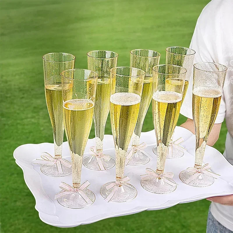 A set of clear plastic champagne flutes with glitter detailing displayed on a white platter outdoors.