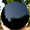 A hand holds a round, glossy black obsidian mirror reflecting surrounding greenery.