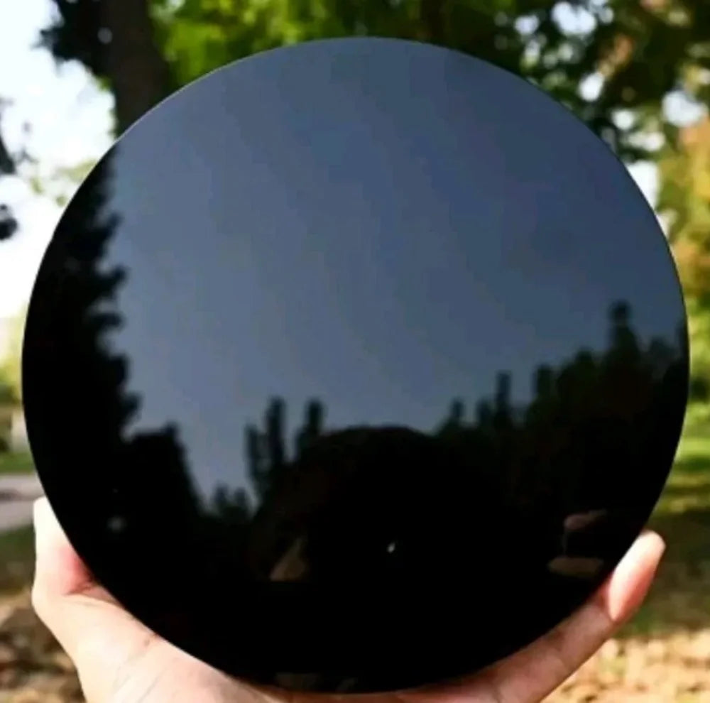 A hand holds a round, glossy black obsidian mirror reflecting surrounding greenery.