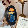 An abstract oval wooden picture frame with vibrant yellow, orange, and blue colors sits next to a gold lamp.