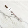 A long, slender metal wand replica with a wooden handle is displayed on a white marble surface.