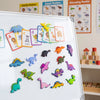 A collection of colorful dinosaur fridge magnets displayed on a white board.