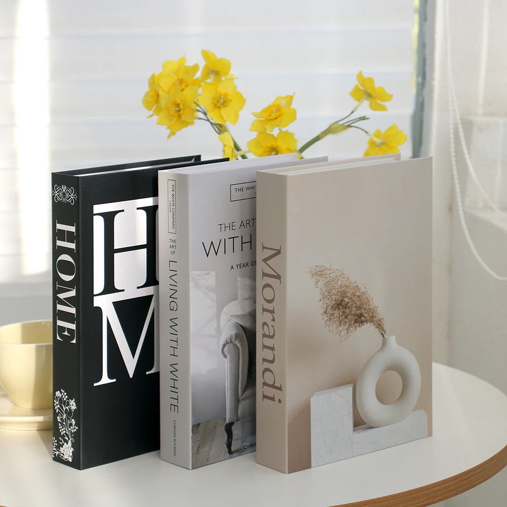 A set of three modern decorative fake books in white and black are displayed next to yellow flowers.