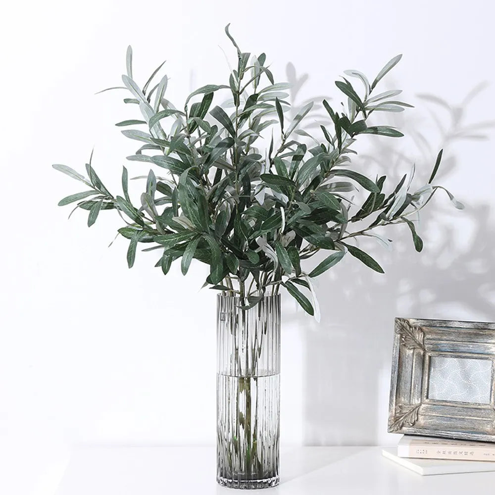 Artificial olive branch with green leaves and small fruits displayed in a clear glass vase.