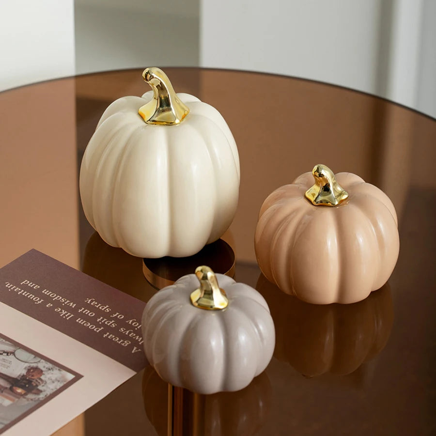 A set of three ceramic pumpkin figurines in cream and blush pink with gold accents displayed on a dark wooden surface.